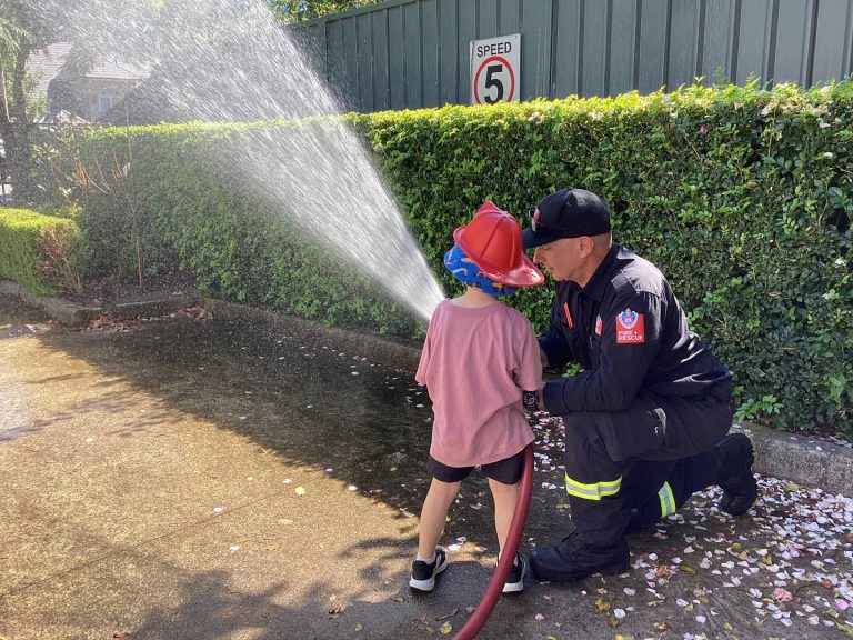 A child in a fire helmet holding the hose with a firefighter during a Fire and Rescue visit