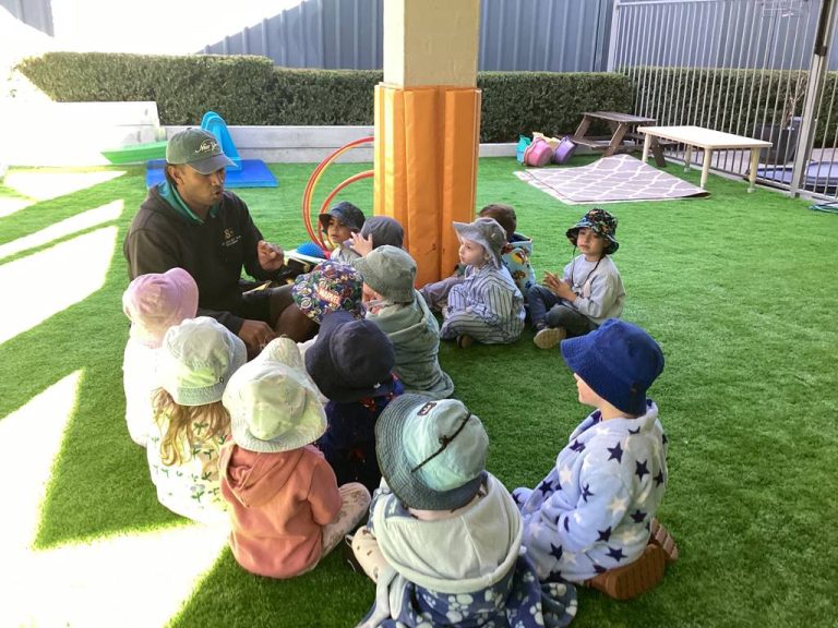An educator reading to a circle of children in the outdoor space at Macarthur Early Learning